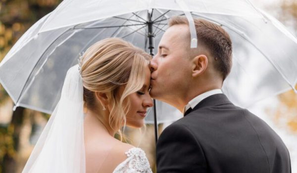 Back view of young groom kisses a blonde bride under an umbrella in the park. Concept of wedding couple